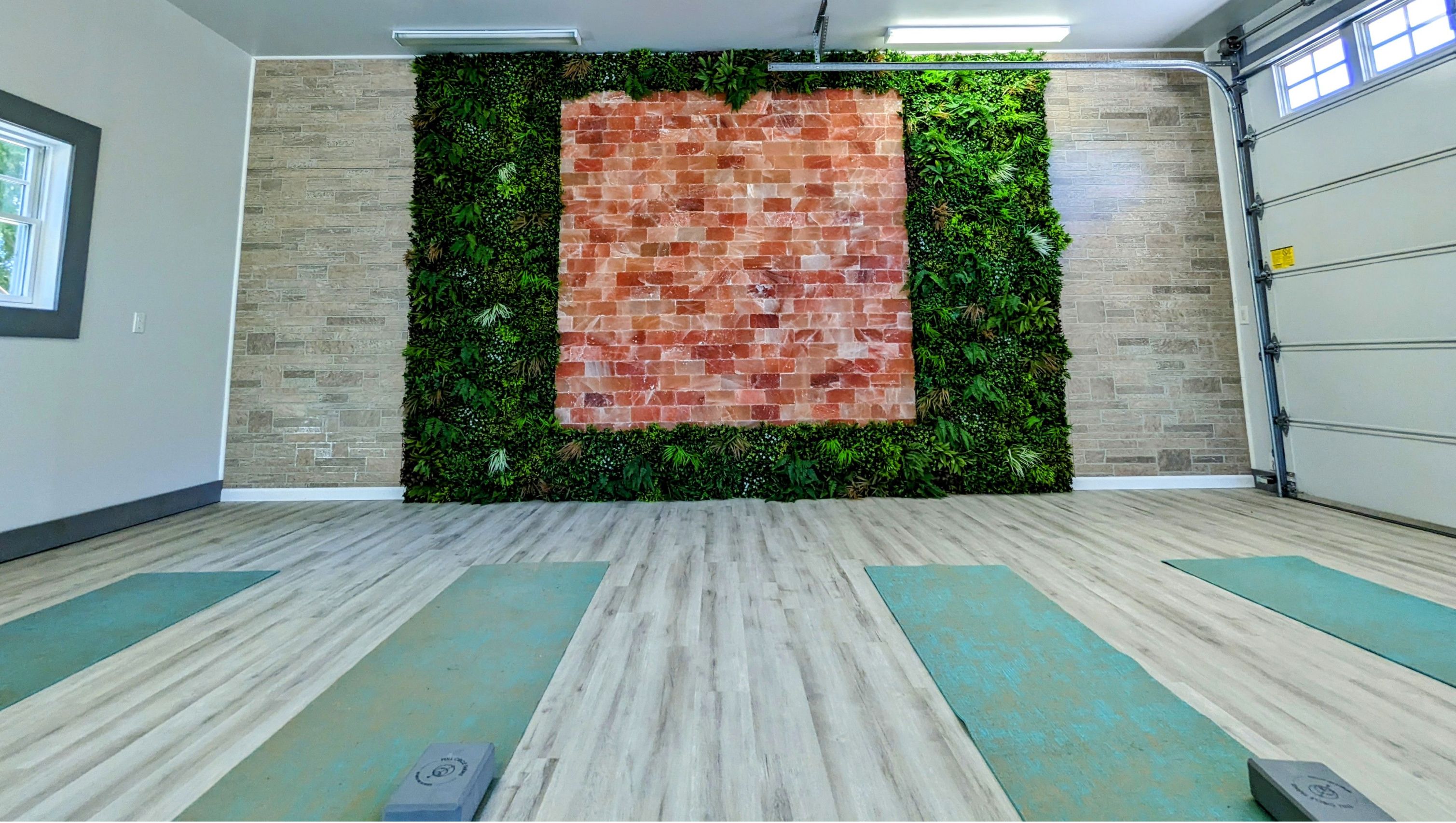 A yoga studio featuring a large wall with pink Himalayan salt bricks framed by vibrant green artificial plants, light wood flooring, and yoga mats.