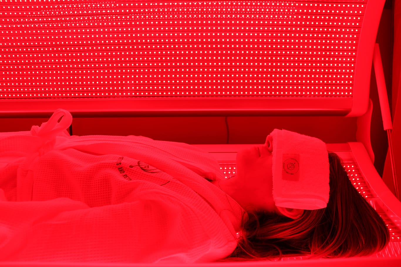Person relaxing in a white robe with the Full Circle Farms logo, a towel over their head, in red light therapy bed