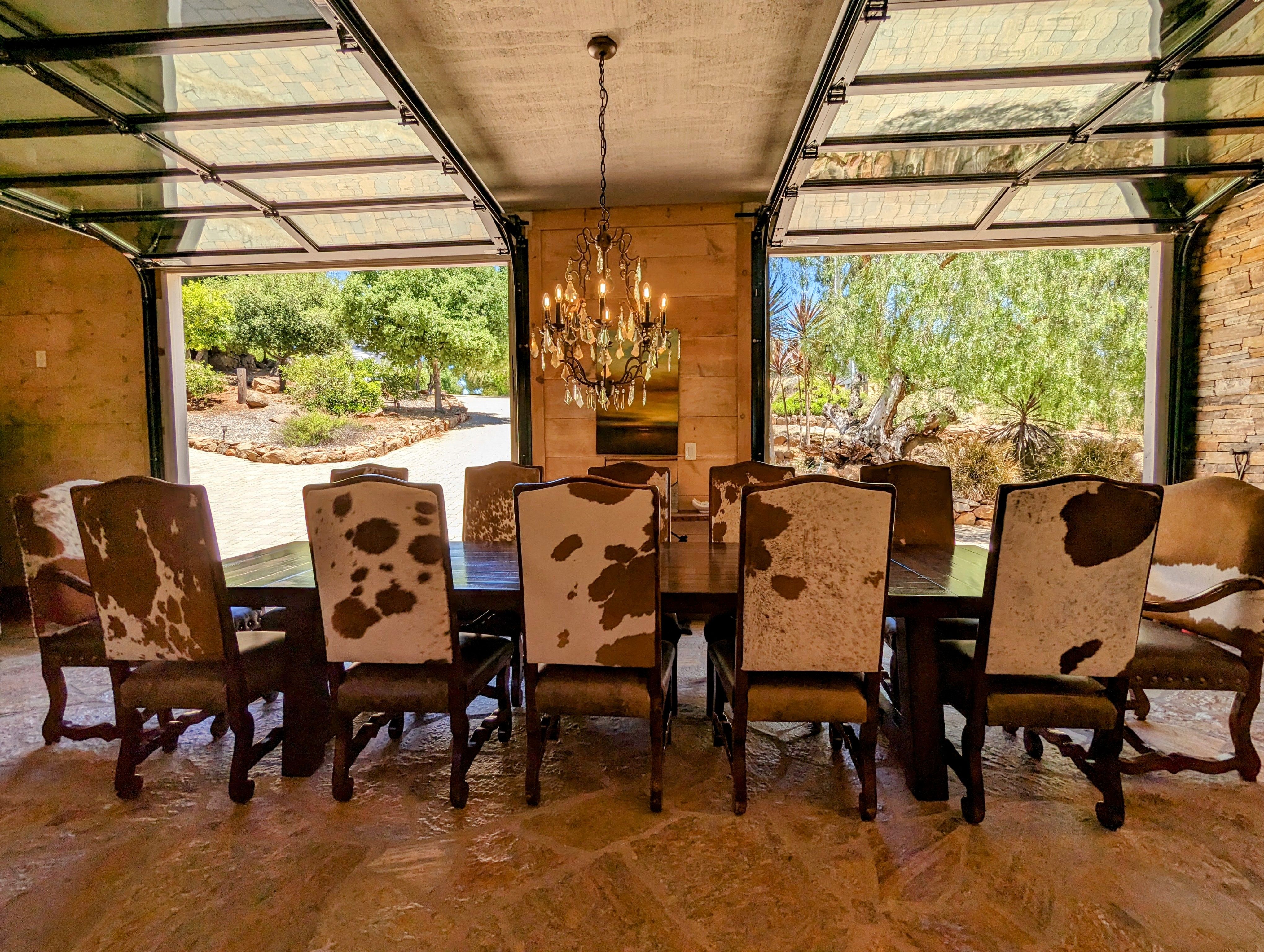 A rustic-modern dining room features a large wooden table with ten cowhide-upholstered chairs. Two glass garage-style doors are open, revealing a sunny, tree-lined outdoor path and landscape. An ornate chandelier hangs from the concrete ceiling.