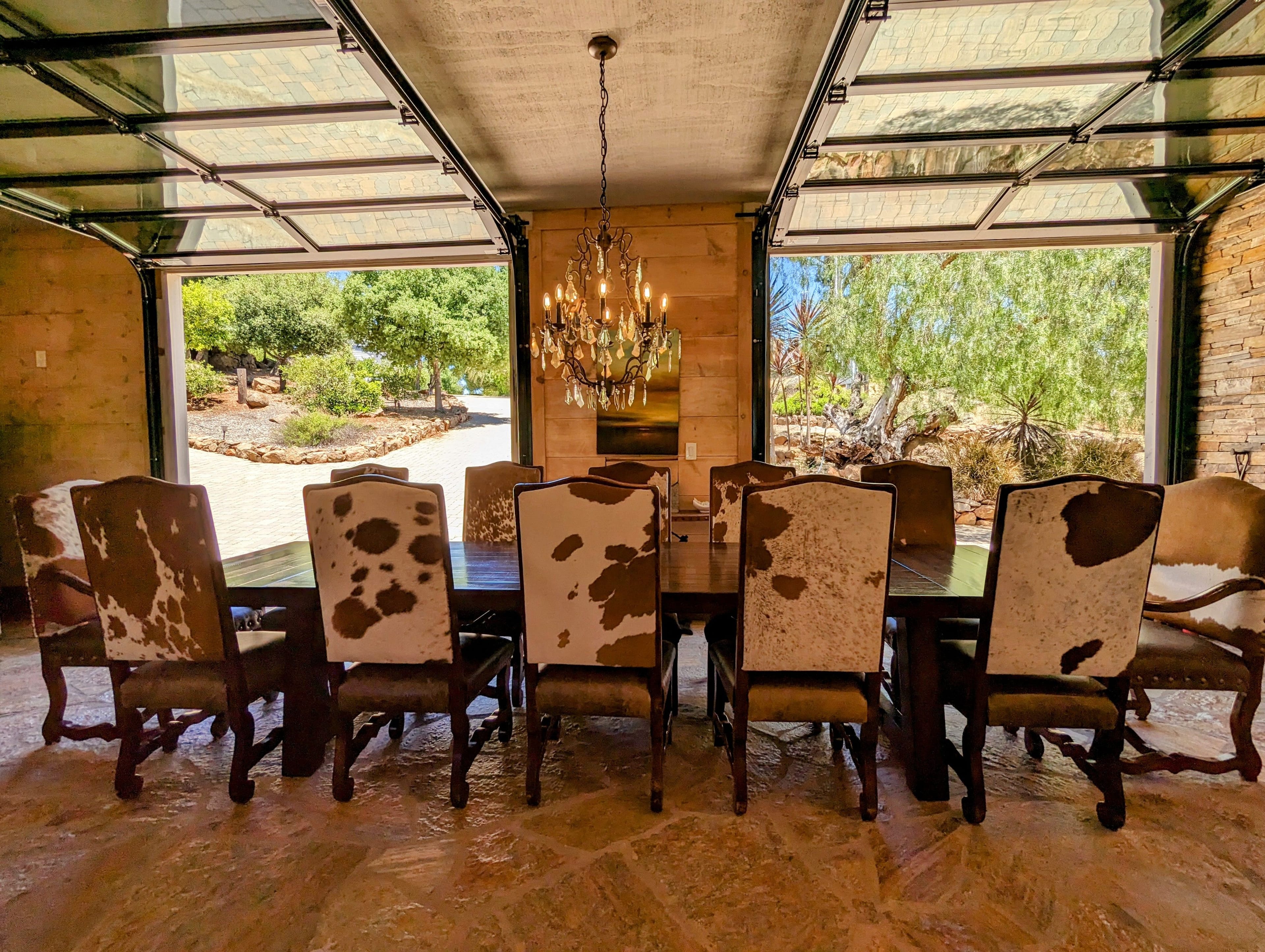Dining room with a long table and cowhide-patterned chairs, featuring overhead glass doors open to a sunny outdoor landscape with trees and a paved path.
