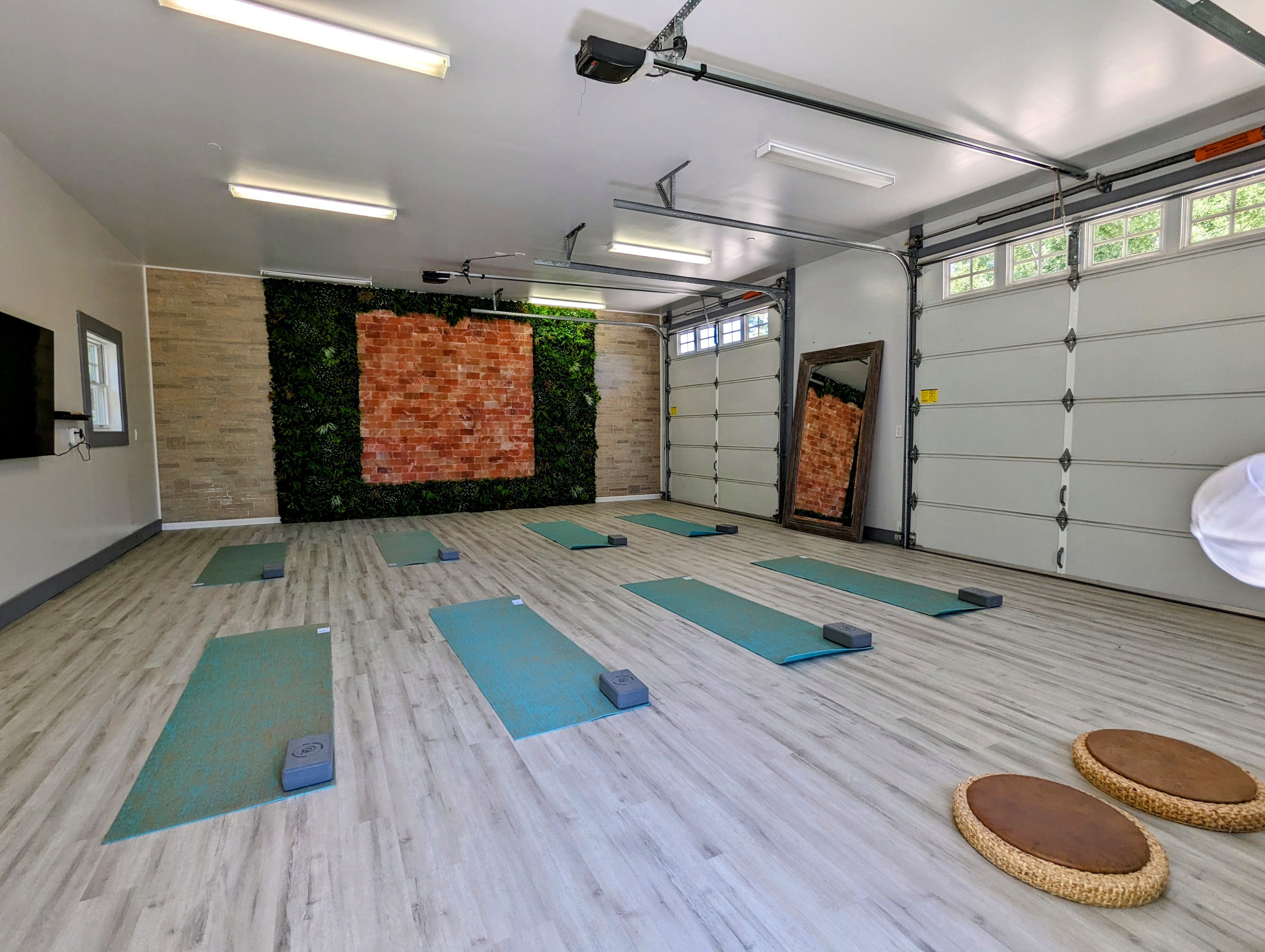 Yoga studio in a repurposed garage featuring light wood floors, several teal yoga mats with blocks, a brick wall accented with artificial greenery, and two large garage doors.