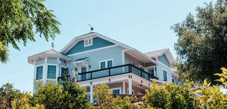 A light blue house with white trim and a dark balcony railing, partially visible through green trees and bushes under a clear blue sky.