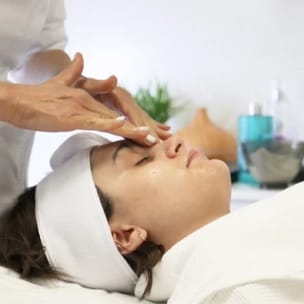 A woman in a white robe and headband lies with her eyes closed as an esthetician massages her forehead during a facial treatment.