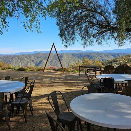Scenic outdoor patio with round white tables and metal chairs under a tree, overlooking rolling green mountains and an A-frame sculpture.