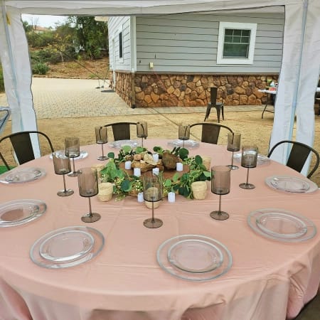 A round table set for an outdoor event, featuring a pink tablecloth, clear plates, dark amber glasses, and a centerpiece of greenery, candles, and rocks under a white tent.