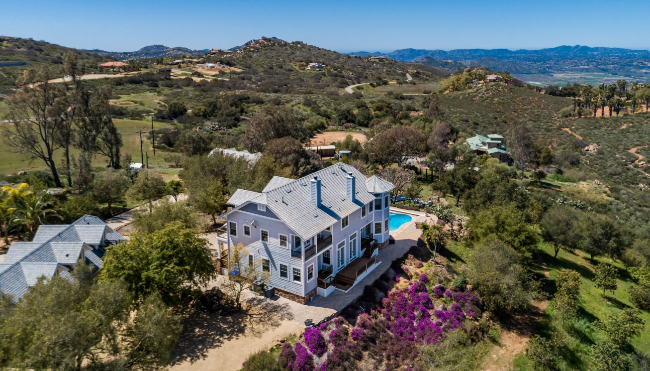 Aerial view of a large light blue house with a swimming pool on a hill, surrounded by green trees, purple flowers, and distant rolling hills under a clear sky.