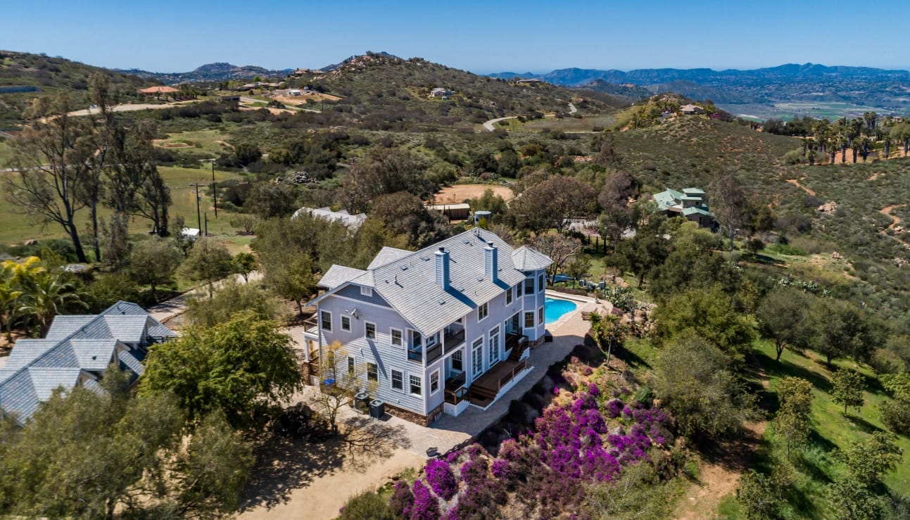 Aerial view of a large light blue house with a swimming pool, situated on a vibrant hillside adorned with green trees and purple flowers, overlooking distant mountains.
