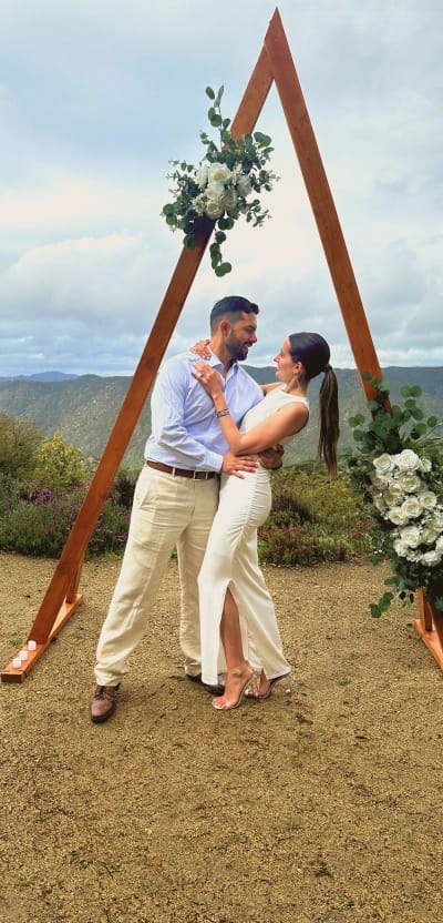 A loving couple embraces under a rustic wooden arch adorned with white flowers and greenery, looking into each other's eyes against a scenic mountain backdrop.