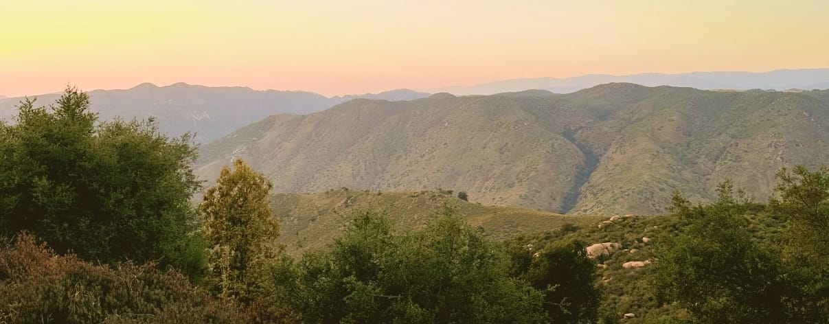 Expansive landscape of green, brush-covered hills and layered mountains, with a prominent valley, beneath a soft yellow and pink sunset sky.
