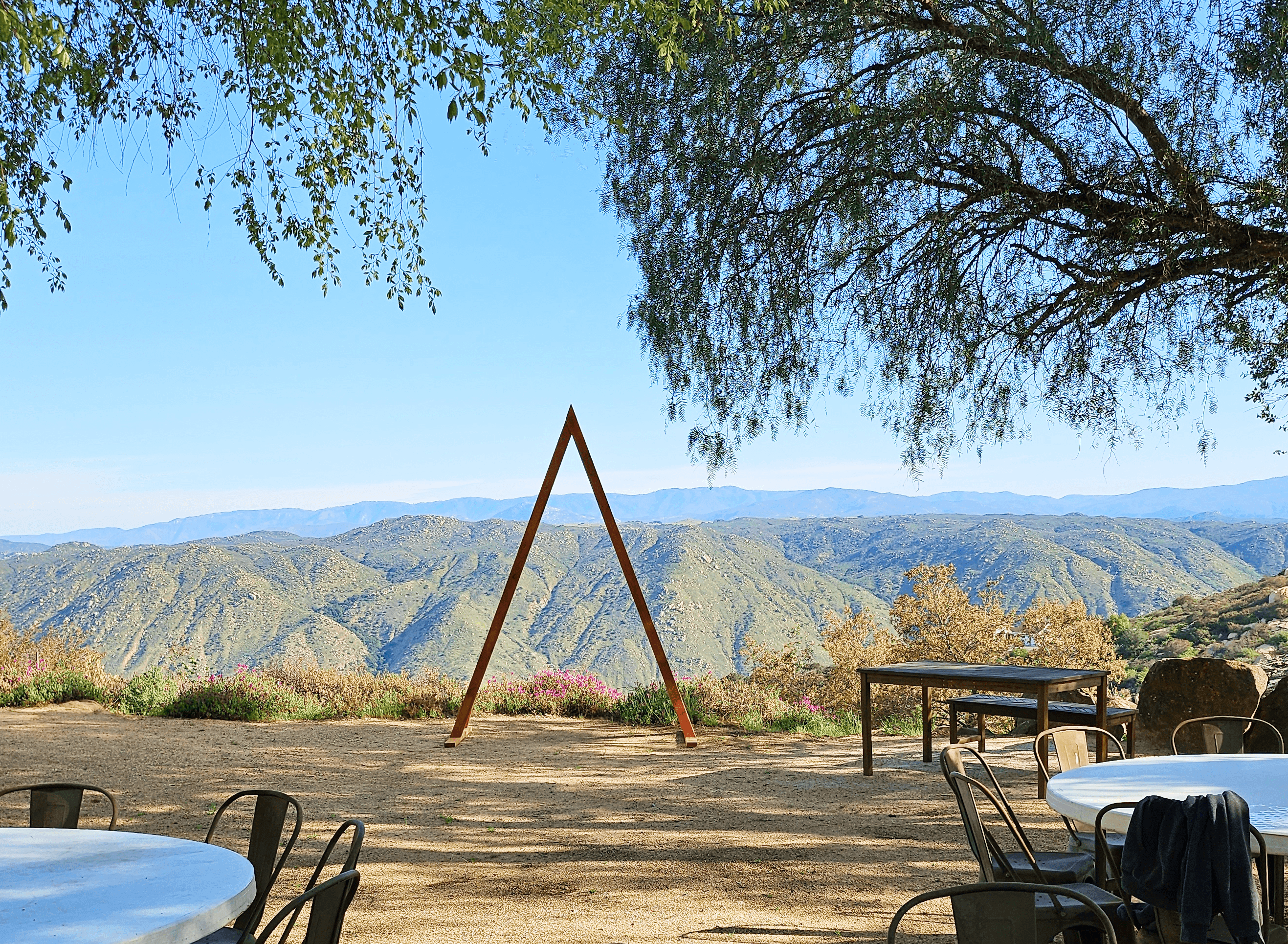 A scenic mountain view from an outdoor patio with tables, chairs, and a distinct A-frame structure. Overhanging green tree branches frame the blue sky.