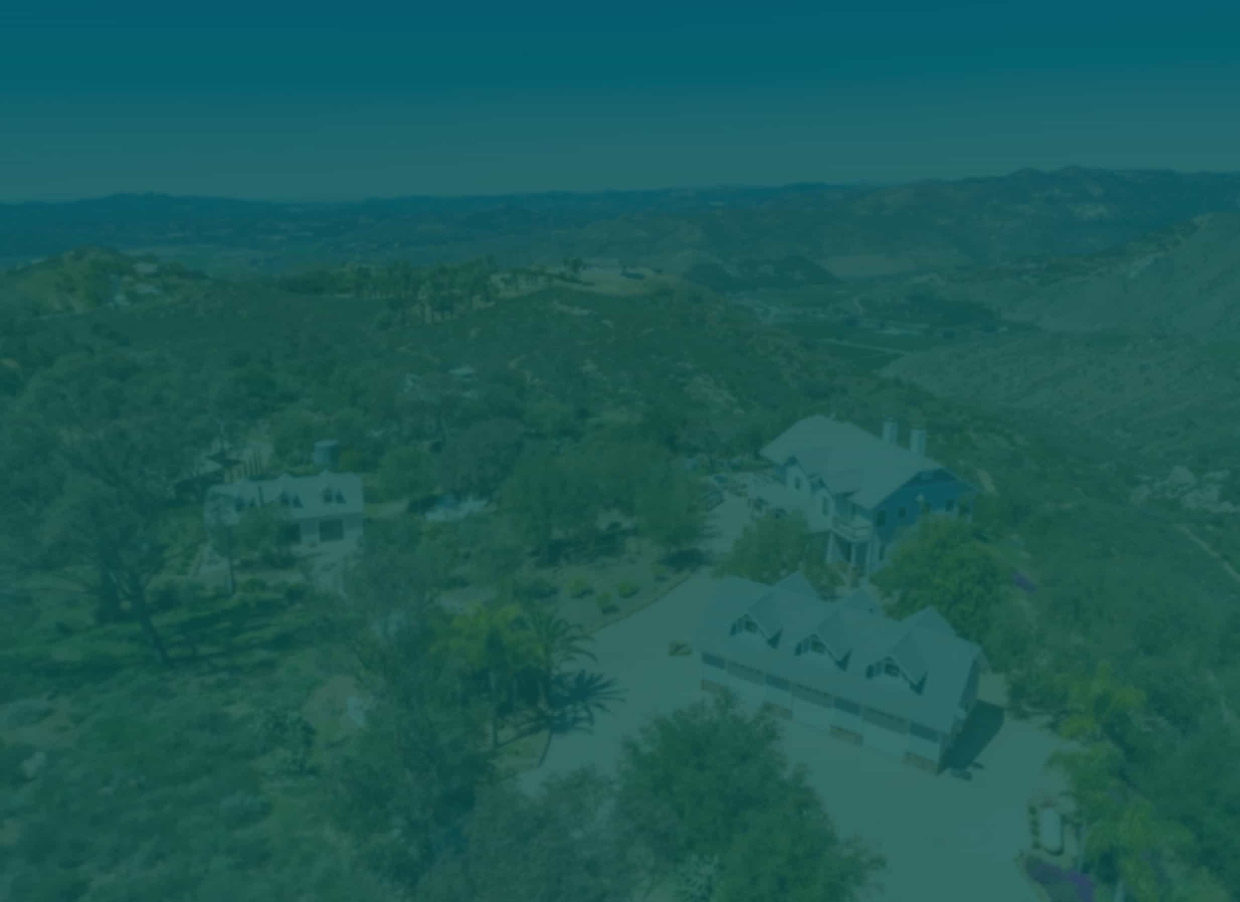 Aerial view of a sprawling estate with multiple large houses nestled among green rolling hills and distant mountains under a clear sky.